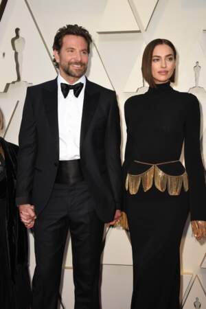 Irina Shayk en robe noire et or Burberry, accompagnée par Bradley Cooper en Tom Ford