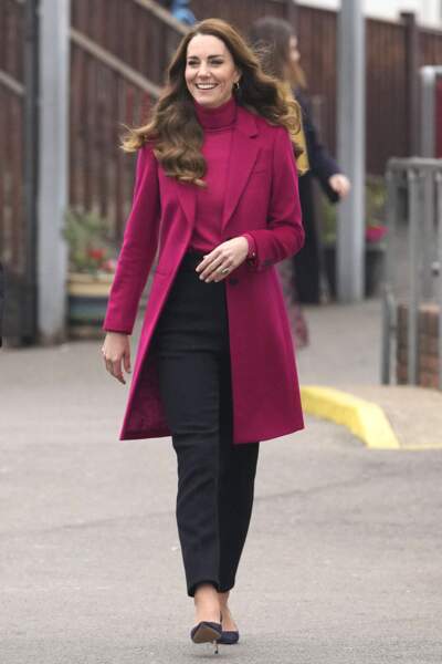 Kate Middleton dans un long manteau fuchsia assorti à son pull col roulé. Elle porte aussi un pantalon noir et des escarpins.