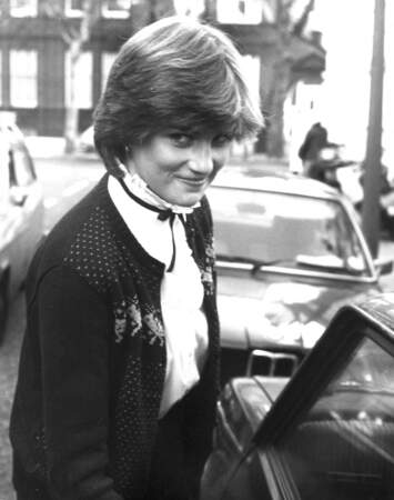 Photographiée dans les rues de Londres à la sortie de son appartement le 15 novembre 1980, Lady Diana arbore un look preppy, composé d'un chemisier blanc à ruban noir, et d'un cardigan en jacquard bordeaux orné de rennes. 