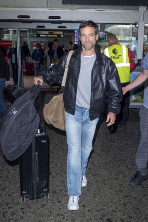 Romain Duris en jean et blouson en cuir aviateur, un sac besace porté à l'épaule et des baskets Salomon.