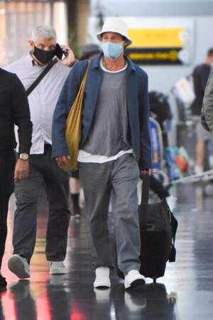 Brad Pitt masqué à l'aéroport de New York en septembre 2021, vêtu d'un pantalon large, d'un bob blanc et d'une veste bleue, un tote bag jaune à son épaule